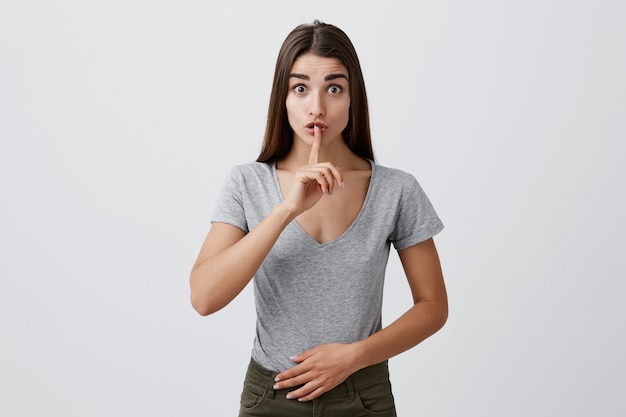 Silencio por favor. Retrato de joven atractiva mujer caucásica encantadora con cabello largo oscuro en ropa casual elegante sosteniendo el dedo índice delante de los labios con las cejas levantadas.