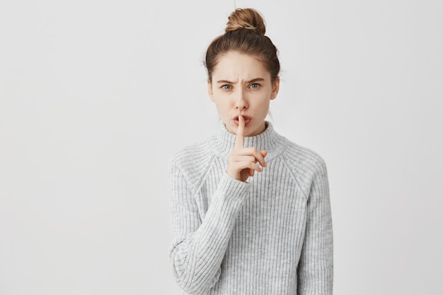 ¡Silencio! Foto de mujer caucásica con dedo índice en los labios. Recepcionista con cabello oscuro atado en moño pidiendo callar diciendo shh sobre la pared blanca. Concepto de silencio