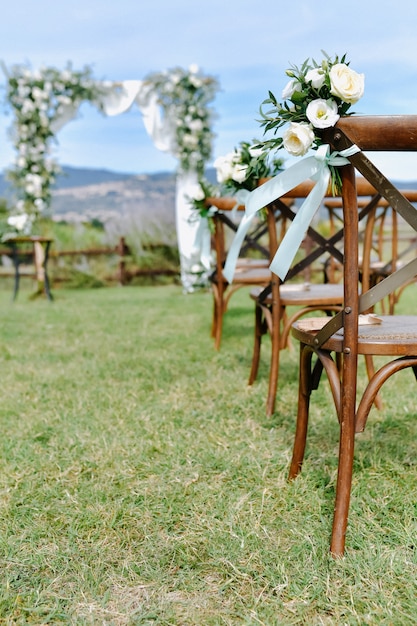 Sillas chiavari marrones decoradas con eustomas blancas en la hierba y el arco de boda decorado en el fondo