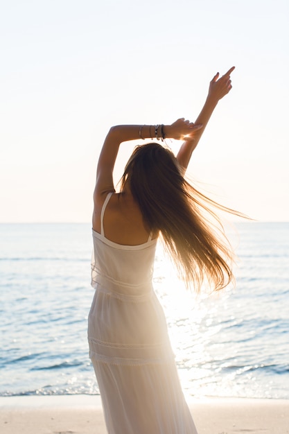 Silueta de una chica delgada de pie en una playa con sol poniente. Lleva un vestido blanco. Tiene el pelo largo que vuela por el aire. Sus brazos se estiraron en el aire