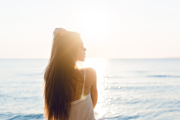 Silueta de una chica delgada de pie en una playa con sol poniente. Lleva un vestido blanco. Tiene el pelo largo que vuela por el aire. Sus brazos se estiraron en el aire