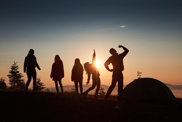 Una silueta de grupo de personas se divierte en la cima de la montaña cerca de la carpa durante la puesta de sol.