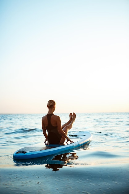Silueta de hermosa joven practicando yoga en tabla de surf en el mar al amanecer.