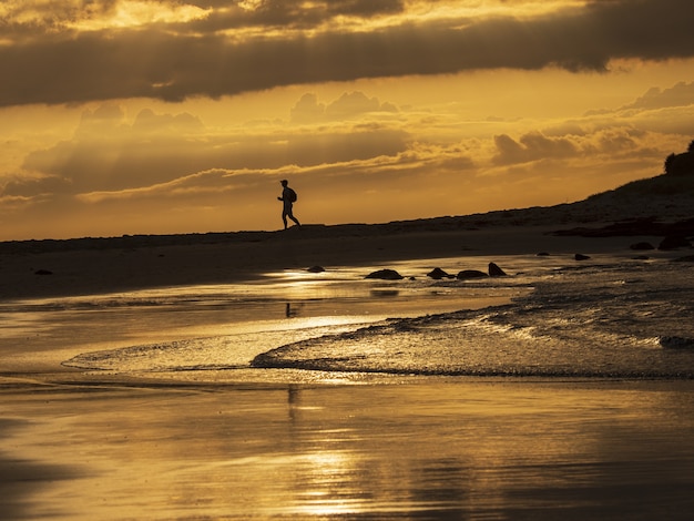 Silueta de un hombre corriendo en la orilla rocosa del mar bajo el cielo dorado del atardecer