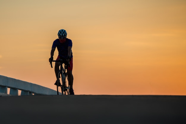 Silueta de un hombre monta una bicicleta al atardecer. Fondo de cielo azul-naranja.