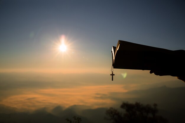 Silueta de la mano humana que sostiene la biblia y la cruz, el fondo es la salida del sol