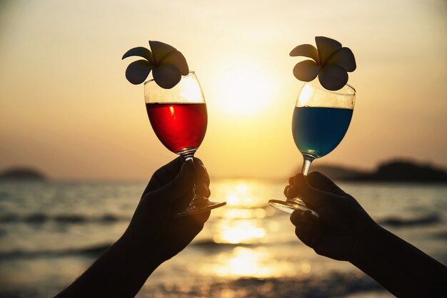 Silueta de la mano de los pares que sostiene la decoración de cristal del cóctel con la flor del plumeria con la playa - feliz relaje las vacaciones de la celebración en concepto del mar