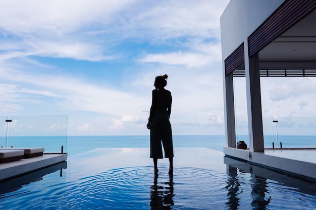 Silueta de una mujer caminando sobre la superficie del agua de la piscina infinita de una costosa villa de lujo rica en una montaña con vista al mar