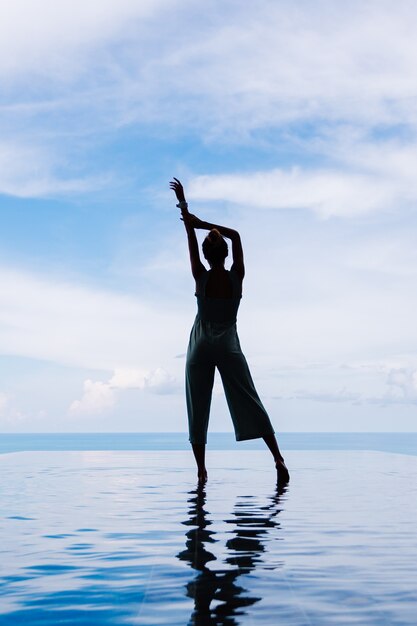 Silueta de una mujer caminando sobre la superficie del agua de la piscina infinita de una costosa villa de lujo rica en una montaña con vista al mar