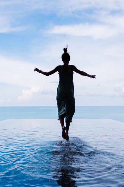 Silueta de una mujer caminando sobre la superficie del agua de la piscina infinita de una costosa villa de lujo rica en una montaña con vista al mar