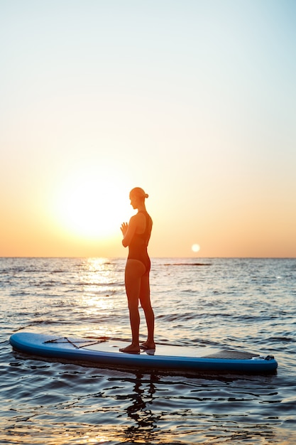 Silueta de mujer hermosa practicando yoga en tabla de surf al amanecer.