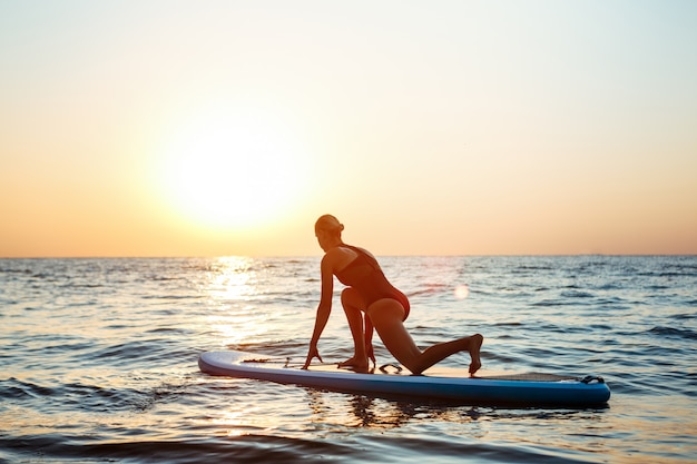 Silueta de mujer hermosa practicando yoga en tabla de surf al amanecer.