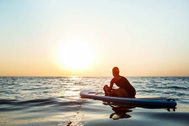 Silueta de mujer hermosa practicando yoga en tabla de surf al amanecer.