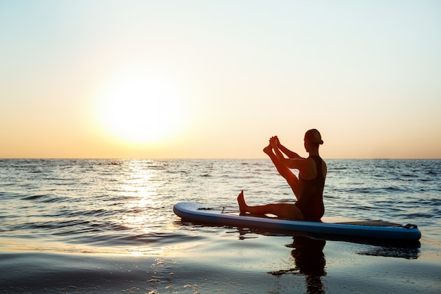 Silueta de mujer hermosa practicando yoga en tabla de surf al amanecer.