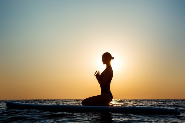 Silueta de mujer hermosa practicando yoga en tabla de surf al amanecer.