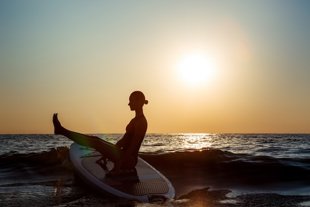 Silueta de mujer hermosa practicando yoga en tabla de surf al amanecer.