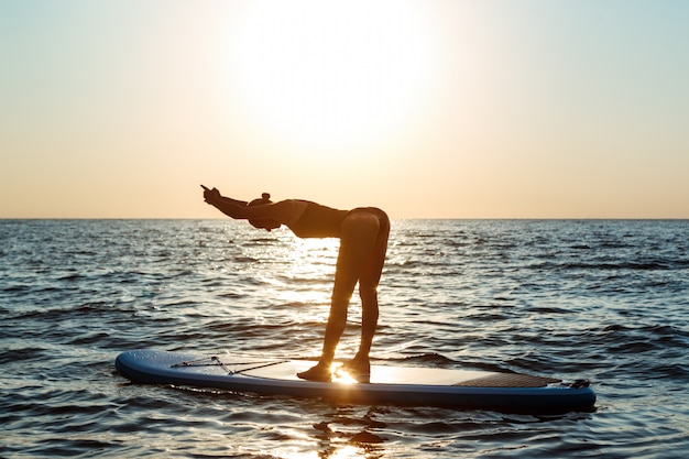 Silueta de mujer hermosa practicando yoga en tabla de surf al amanecer.