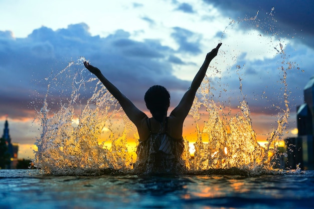 Silueta negra de mujer asiática salpicando agua en vacaciones de verano relajándose en una piscina infinita con vistas al mar azul al atardecer con rascacielos de gran altura en el centro urbano Estilo de vida de felicidad saludable