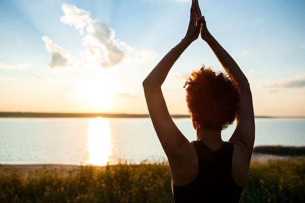 Silueta de niña deportiva practicando yoga en el campo al amanecer.