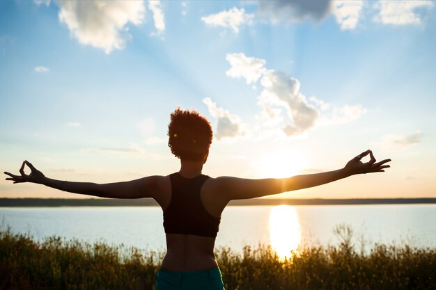 Silueta de niña deportiva practicando yoga en el campo al amanecer.