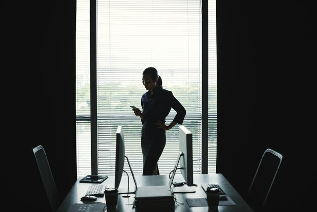 Silueta oscura de mujer de pie contra la ventana en la oficina y usando el teléfono inteligente