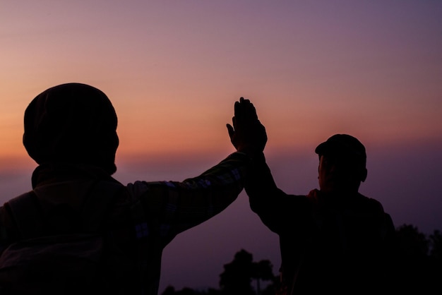 Silueta de trabajo en equipo ayudando a la confianza de la mano ayuda Éxito en las montañas Los excursionistas celebran con las manos en alto Ayúdense unos a otros en la cima de la montaña y el paisaje de la puesta del sol