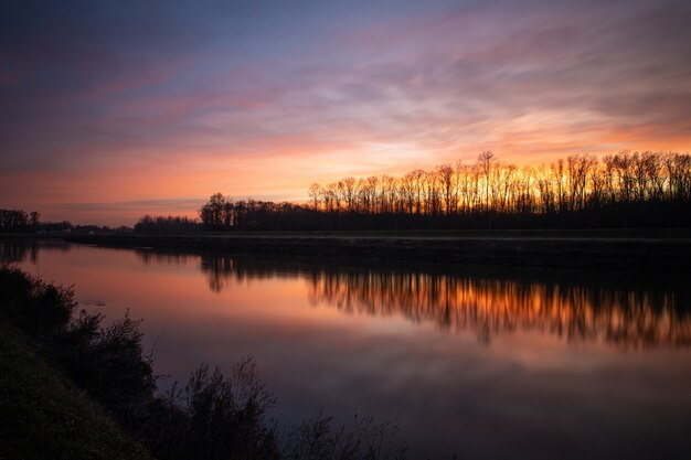 Siluetas de árboles bajo el cielo nublado del atardecer reflejado en el lago de abajo