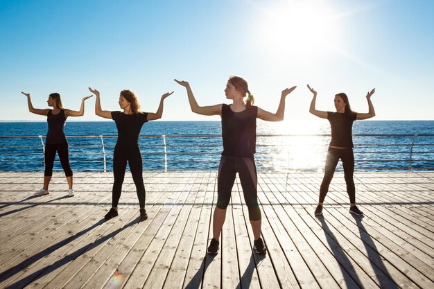 Siluetas de chicas deportivas bailando cerca del mar al amanecer.