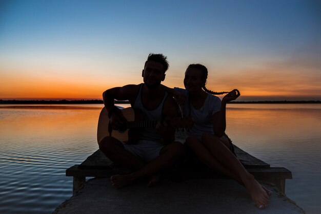 Siluetas de joven pareja hermosa descansando regocijo al amanecer cerca del lago