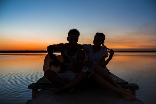 Siluetas de joven pareja hermosa descansando regocijo al amanecer cerca del lago