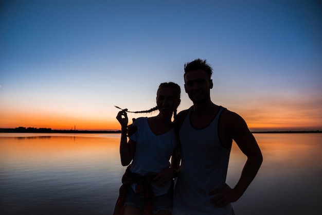 Siluetas de joven pareja hermosa descansando regocijo al amanecer cerca del lago