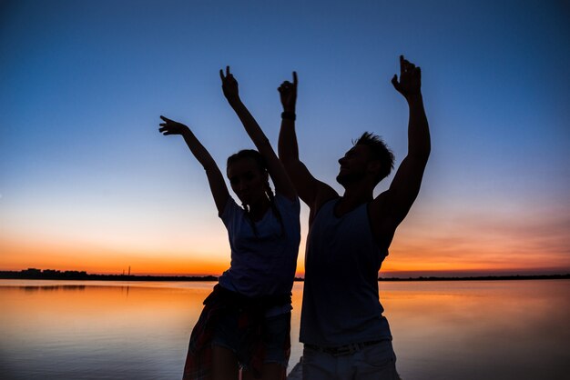 Siluetas de joven pareja hermosa descansando regocijo al amanecer cerca del lago