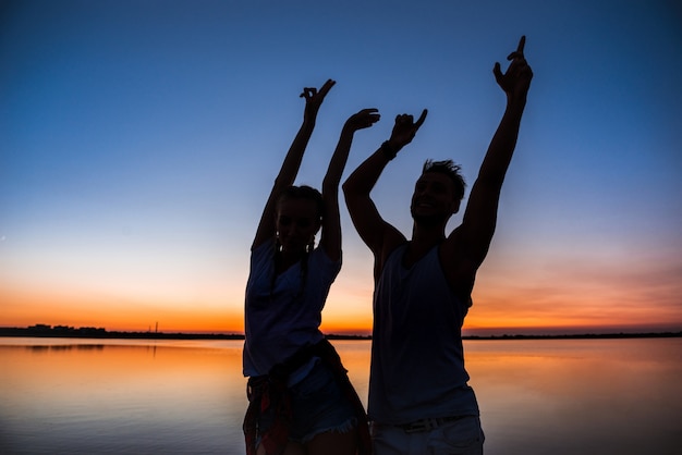 Siluetas de joven pareja hermosa descansando regocijo al amanecer cerca del lago