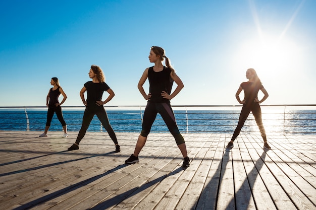 Siluetas de mujeres deportivas bailando zumba cerca del mar al amanecer