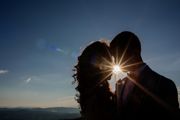 Siluetas de novios de pie en los rayos del sol antes del paisaje de montaña