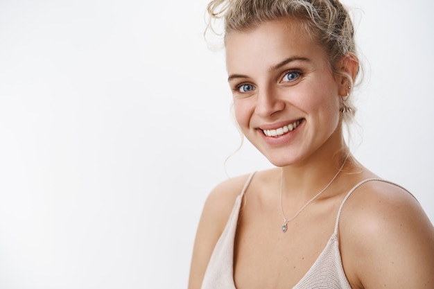 Sincera y encantada chica rubia femenina con ojos azules y una agradable sonrisa feliz mirando a la cámara riendo divirtiéndose, riendo disfrutando de la noche, posando satisfecha y alegre en pose tierna sobre una pared blanca