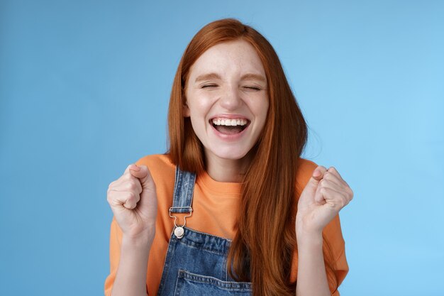 Sincero feliz regocijo chica jengibre cerrar los ojos sonriendo ampliamente decir sí agitando los puños cerrados con alegría ...