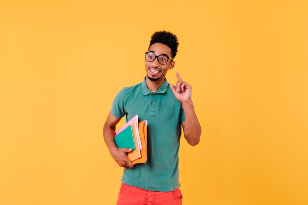 Situación inteligente de estudiante internacional. Foto de hombre africano extático lleva gafas elegantes con libros.