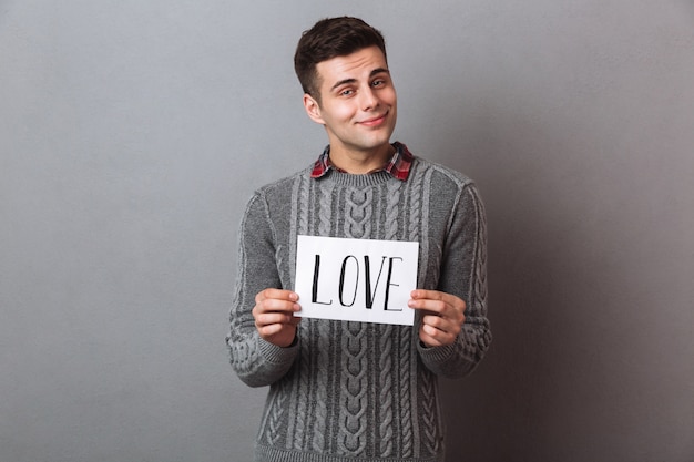 Situación sonriente joven del hombre aislada sosteniendo el papel con el texto del amor.