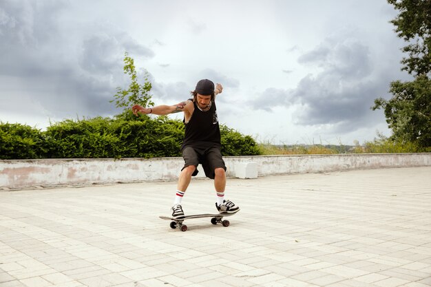 Skater haciendo un truco en la calle de la ciudad en un día nublado. Hombre joven en zapatillas y gorra de montar y hacer longboard sobre el asfalto. Concepto de actividad de ocio, deporte, extremo, afición y movimiento.