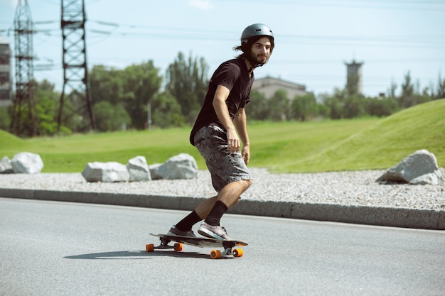 Skater haciendo un truco en la calle de la ciudad en un día soleado. Hombre joven en equipo de equitación y longboard sobre el asfalto en acción. Concepto de actividad de ocio, deporte, extremo, afición y movimiento.