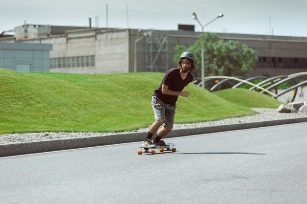 Skater haciendo un truco en la calle de la ciudad en un día soleado. Hombre joven en equipo de equitación y longboard sobre el asfalto en acción. Concepto de actividad de ocio, deporte, extremo, afición y movimiento.