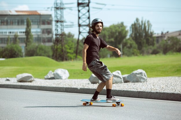 Skater haciendo un truco en la calle de la ciudad en un día soleado. Hombre joven en equipo de equitación y longboard sobre el asfalto en acción. Concepto de actividad de ocio, deporte, extremo, afición y movimiento.