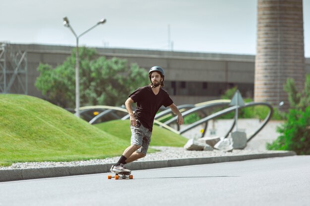 Skater haciendo un truco en la calle de la ciudad en un día soleado. Hombre joven en equipo de equitación y longboard sobre el asfalto en acción. Concepto de actividad de ocio, deporte, extremo, afición y movimiento.