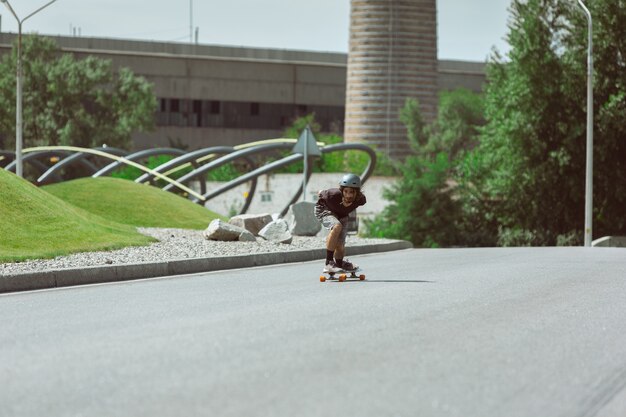 Skater haciendo un truco en la calle de la ciudad en un día soleado. Hombre joven en equipo de equitación y longboard sobre el asfalto en acción. Concepto de actividad de ocio, deporte, extremo, afición y movimiento.