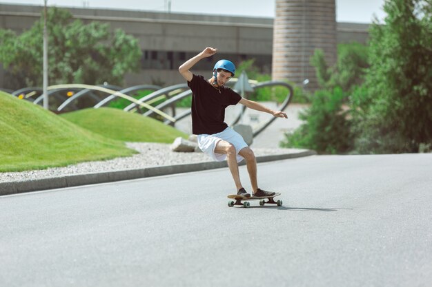 Skater haciendo un truco en la calle de la ciudad en un día soleado. Hombre joven en equipo de equitación y longboard sobre el asfalto en acción. Concepto de actividad de ocio, deporte, extremo, afición y movimiento.