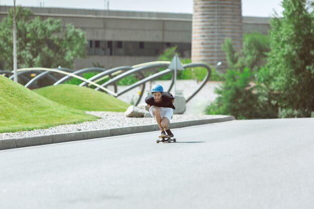 Skater haciendo un truco en la calle de la ciudad en un día soleado. Hombre joven en equipo de equitación y longboard sobre el asfalto en acción. Concepto de actividad de ocio, deporte, extremo, afición y movimiento.