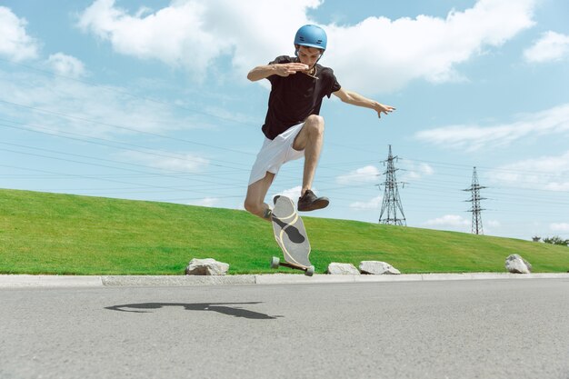 Skater haciendo un truco en la calle de la ciudad en un día soleado. Joven en equipo de equitación y longboard cerca de pradera en acción. Concepto de actividad de ocio, deporte, extremo, afición y movimiento.