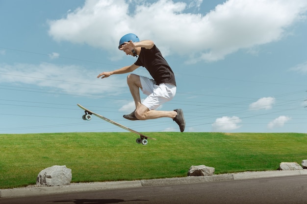 Skater haciendo un truco en la calle de la ciudad en un día soleado. Joven en equipo de equitación y longboard cerca de pradera en acción. Concepto de actividad de ocio, deporte, extremo, afición y movimiento.