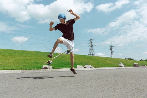 Skater haciendo un truco en la calle de la ciudad en un día soleado. Joven en equipo de equitación y longboard cerca de pradera en acción. Concepto de actividad de ocio, deporte, extremo, afición y movimiento.